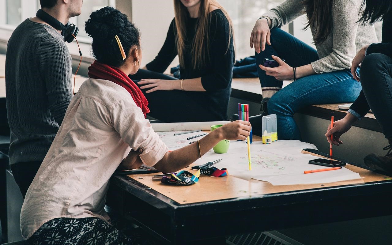 Studenci siedzą wokół stolika, na którym leżą kartki i kolorowe długopisy.