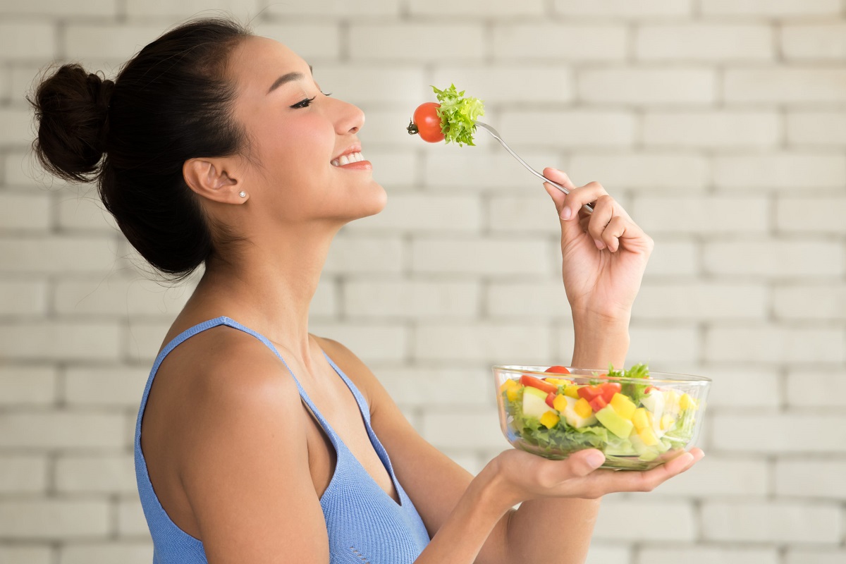 Młoda kobieta uśmiecha się w jednej ręce trzyma naczynie z sałatkę ze świeżych warzyw a w drugiej ręce widelec na który nabity jest liść sałaty i pomidor koktajlowy