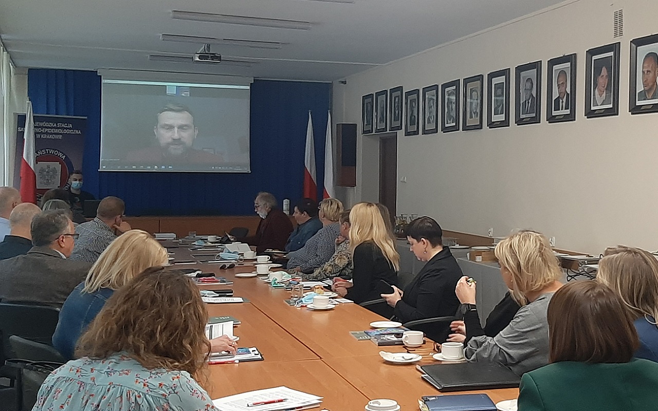 Przy długim stole siedzą uczestnicy spotkania. Ich oczy zwrócone są ku dużemu ekranowi na środku sali , na którym wyświetlany jest obraz przemawiającego prelegenta