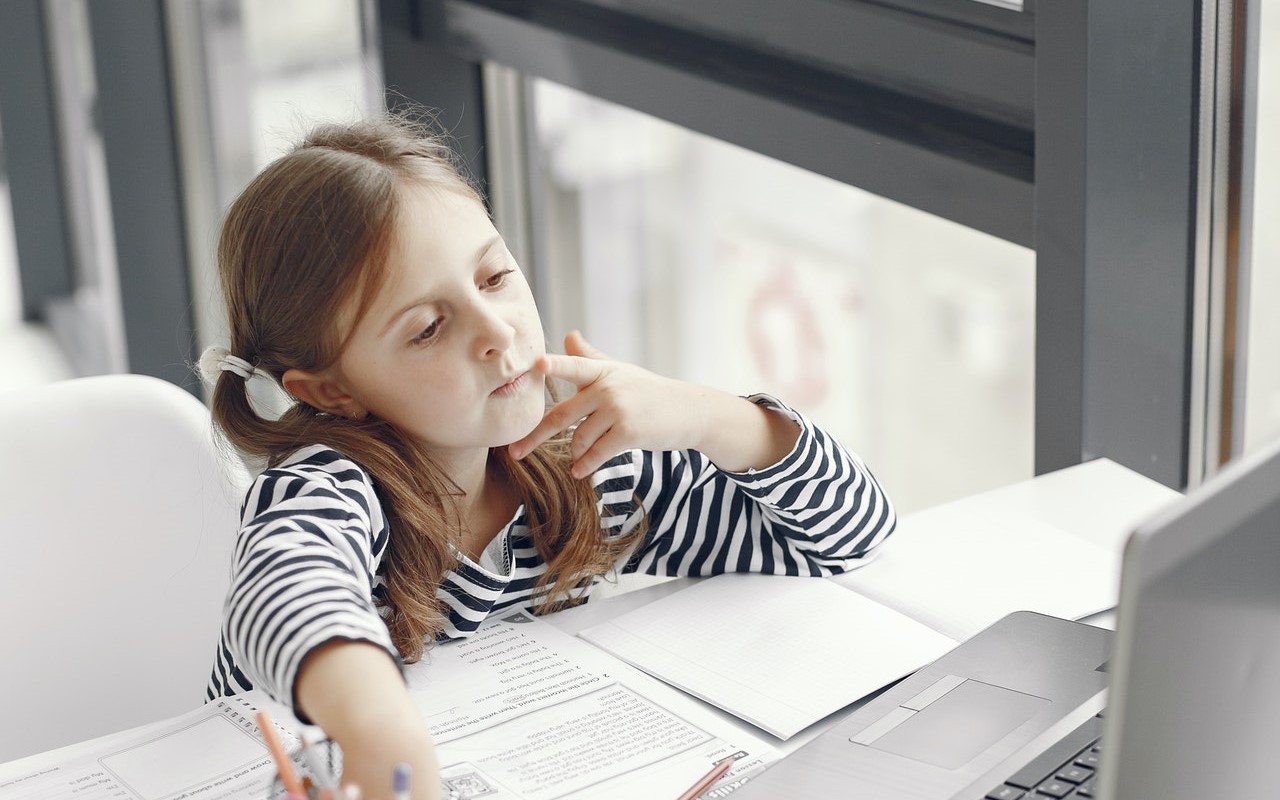 Kilkuletnia dziewczynka siedzi przy biurku patrząc z zastanowieniem w ekran laptopa. Przed nią leżą rozłożone zeszyty i książki.