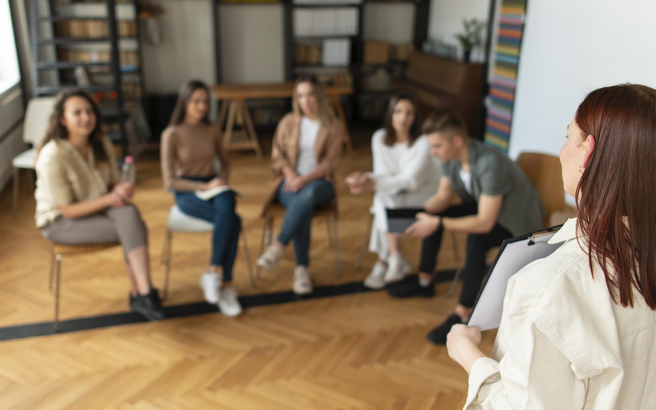 grupa młodych ludzi siedzi w pomieszczeniu wypełnionym książkami i słucha stojącej przed nimi kobiety